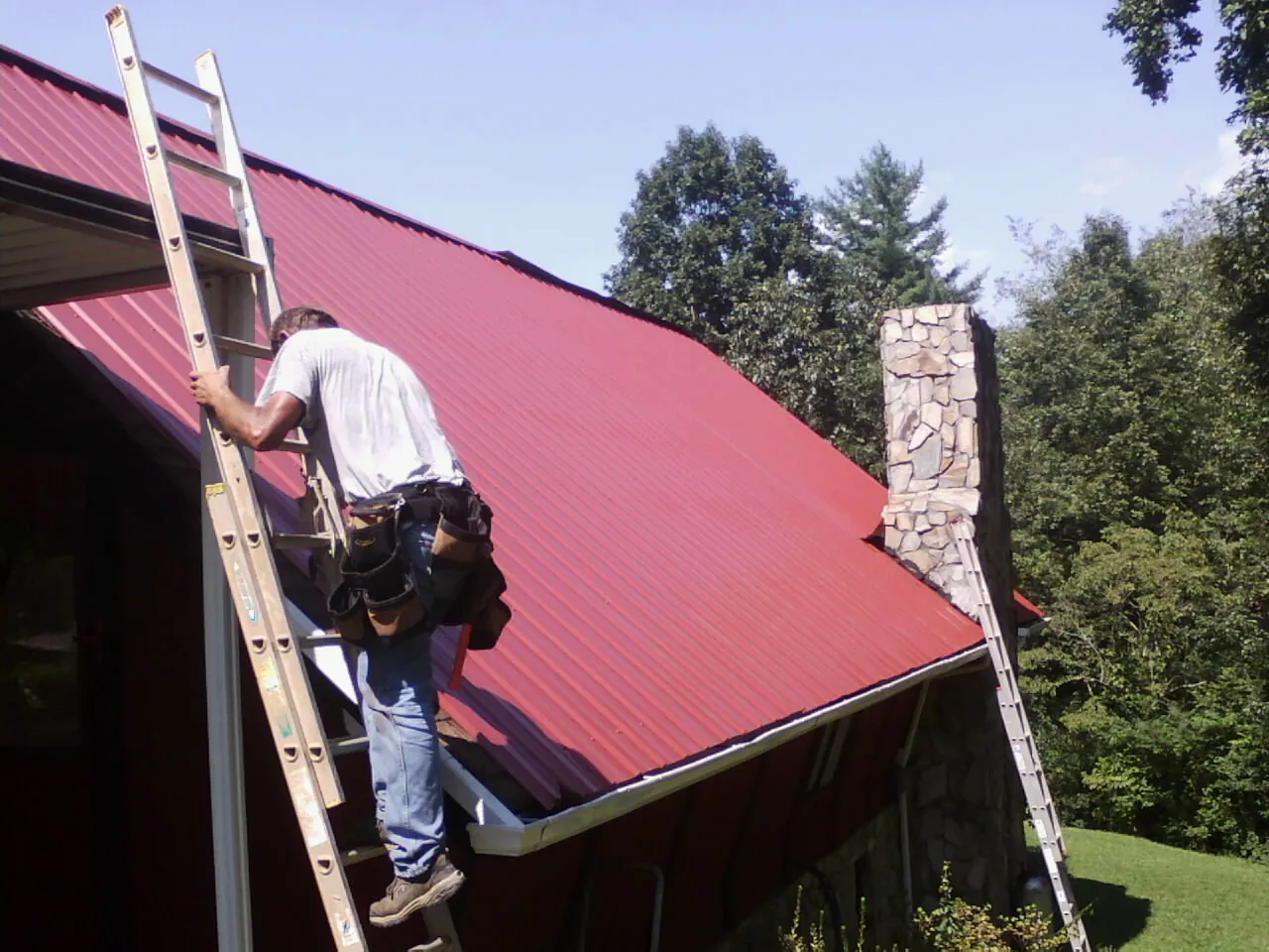 Roofer on a ladder beside a red metal roof and stone chimney — Soffit & Fascia Repair by Roofing Brothers in Fair Lawn